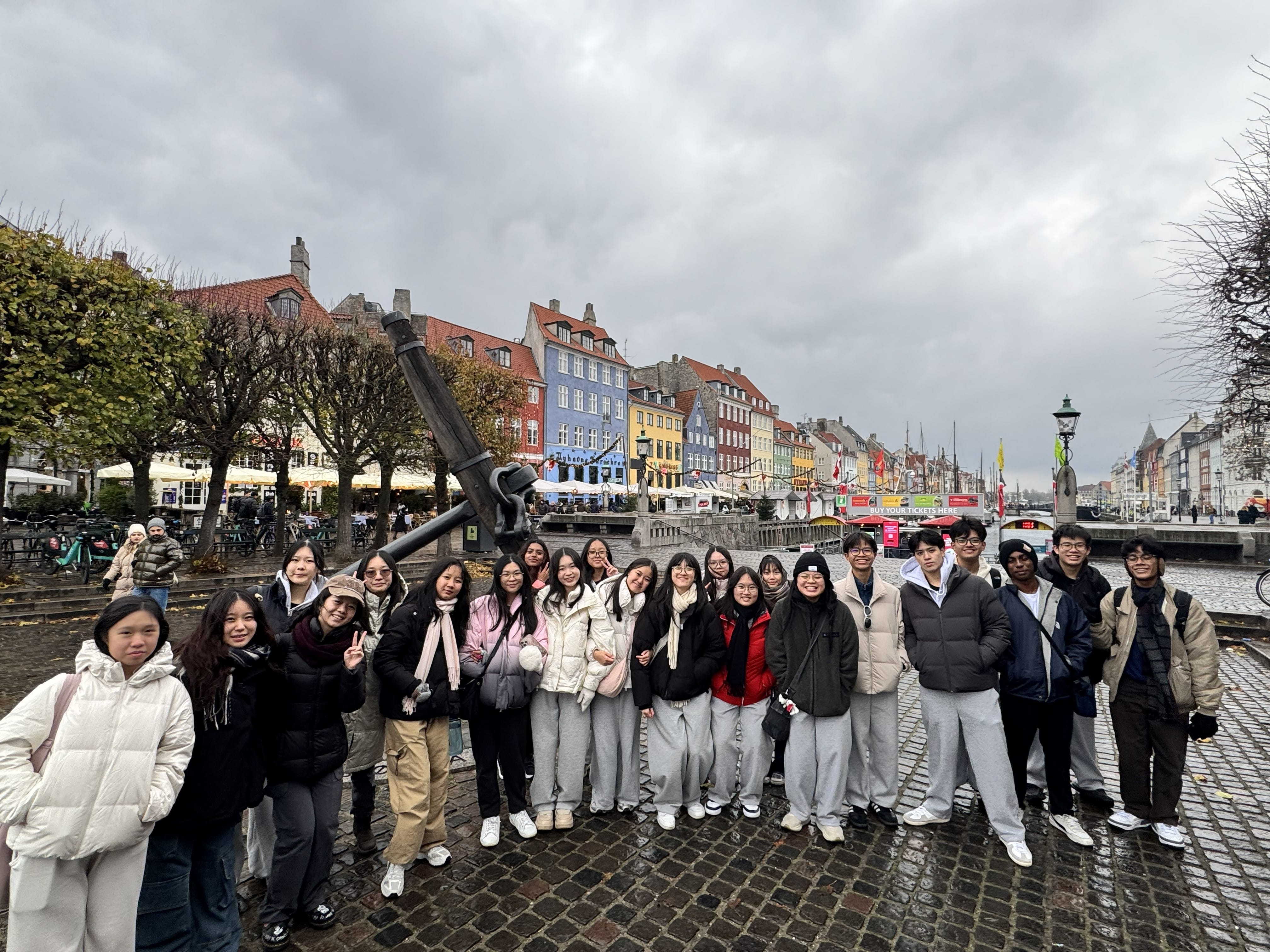 About 25 students in winter clothing pose together on the cobblestone waterfront of Nyhavn, Copenhagen. The iconic colourful canal-side buildings and a large black anchor monument are visible behind them on a cloudy day.criptive alt text.